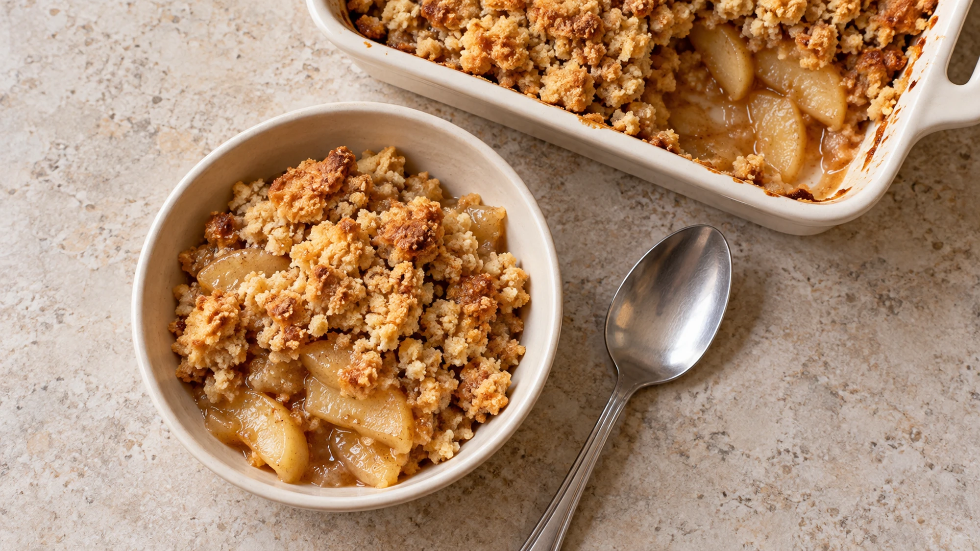 A bowl of apple crumble showing cinnamon apples and a crisp golden topping, with the baking dish behind it.