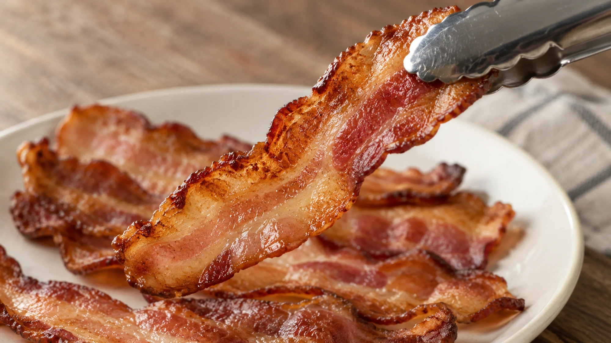 Tongs lifting a crisp strip of thick-cut oven bacon.