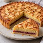 A whole almond-topped Bakewell tart with a separate slice on a round plate.