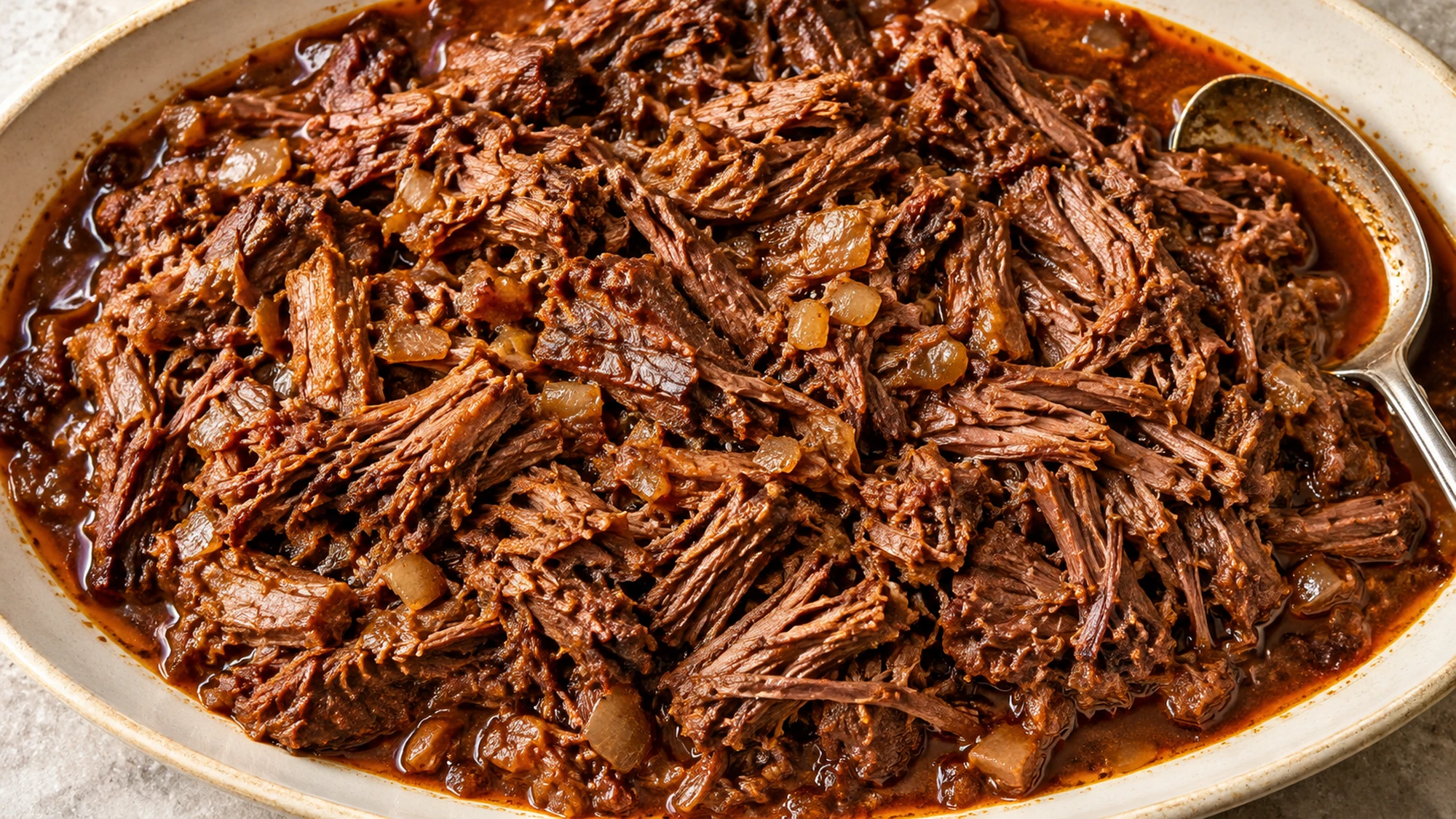 Overhead view of shredded barbacoa with softened onion and chipotle juices on a platter.