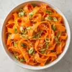 Square overhead view of carrot ribbon salad with scallions and sesame in a cream bowl.