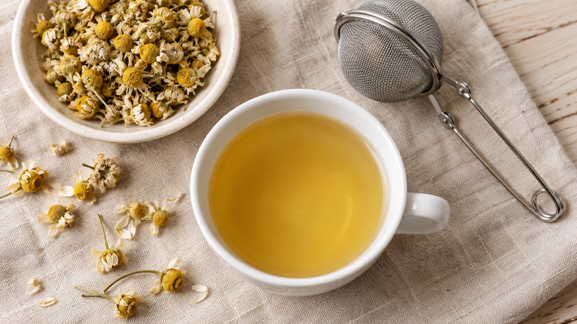 White cup of chamomile tea beside dried flowers and a mesh infuser