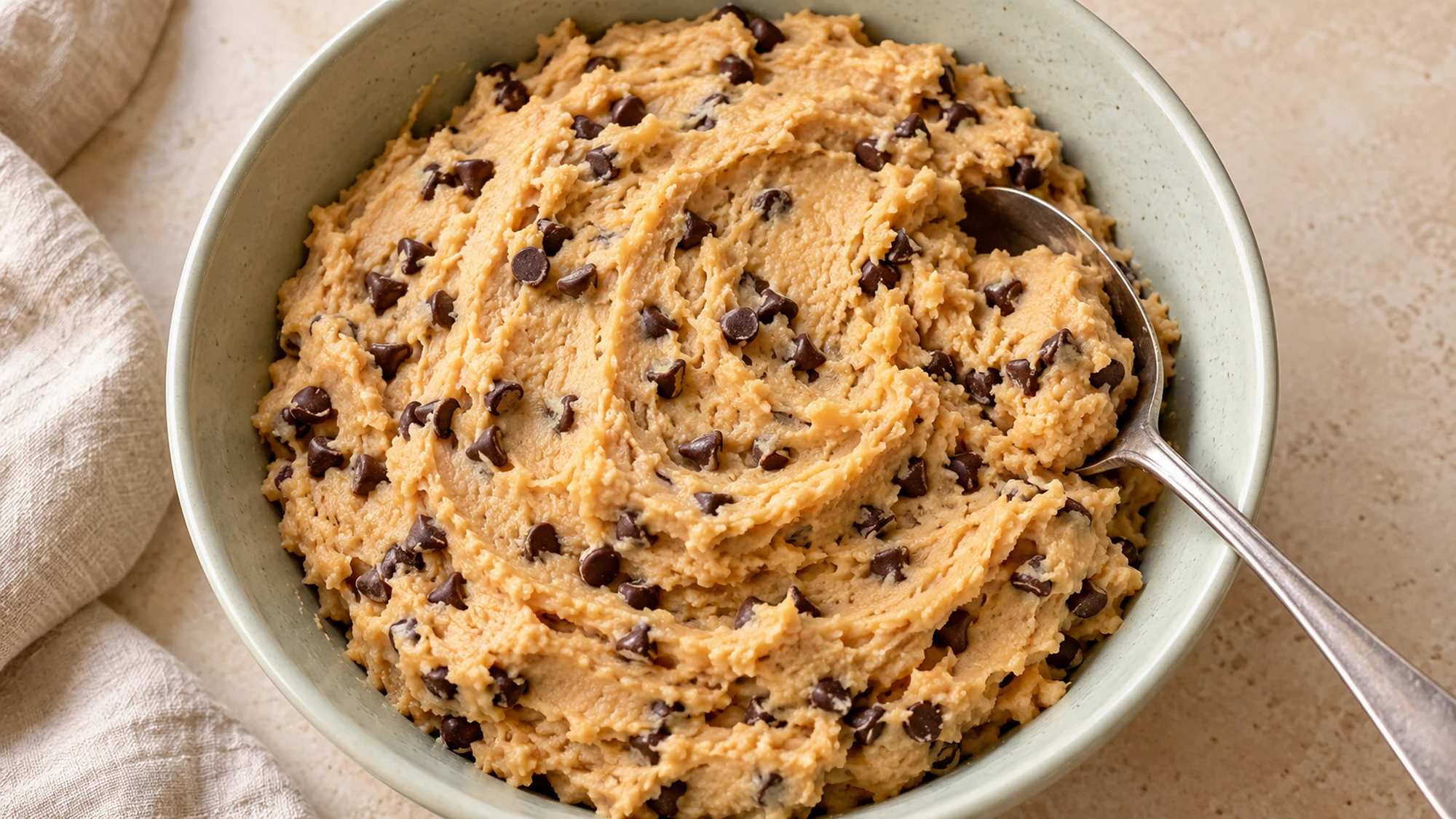 Overhead view of smooth chickpea cookie dough dotted with mini chocolate chips in a sage bowl.