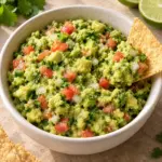 Square close-up of chunky guacamole with a tortilla chip beside the bowl