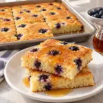 Square view of sheet-pan pancakes showing tender pale-golden pancake squares with blueberries and a sliced cut edge