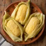 Three pale yellow fresh-corn uchepos arranged in their husks on a terracotta plate.