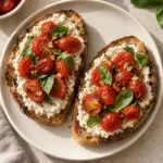 Overhead view of two tomato-topped vegan ricotta toasts on a cream plate.